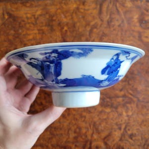 May include: A white and blue porcelain bowl with a flared rim and a short, cylindrical base. The bowl features a blue landscape design around the exterior. The bowl is held by a hand.