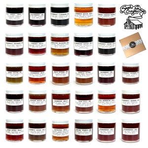 May include: A collection of 30 small glass jars filled with various jams and jellies. The jars have white lids and labels with the names of the flavors printed in black. The jars are arranged in five rows of six jars each. The jars are all the same size and shape. The jars are on a white background. The text on the labels reads: "Smoke Camp Crafts".