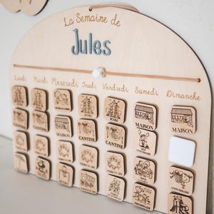 May include: Wooden weekly schedule board with the text "La Semaine de Jules". The board has a series of small wooden tiles with engraved illustrations and text, including "CANTINE" and "MAISON". The days of the week are labeled across the top.