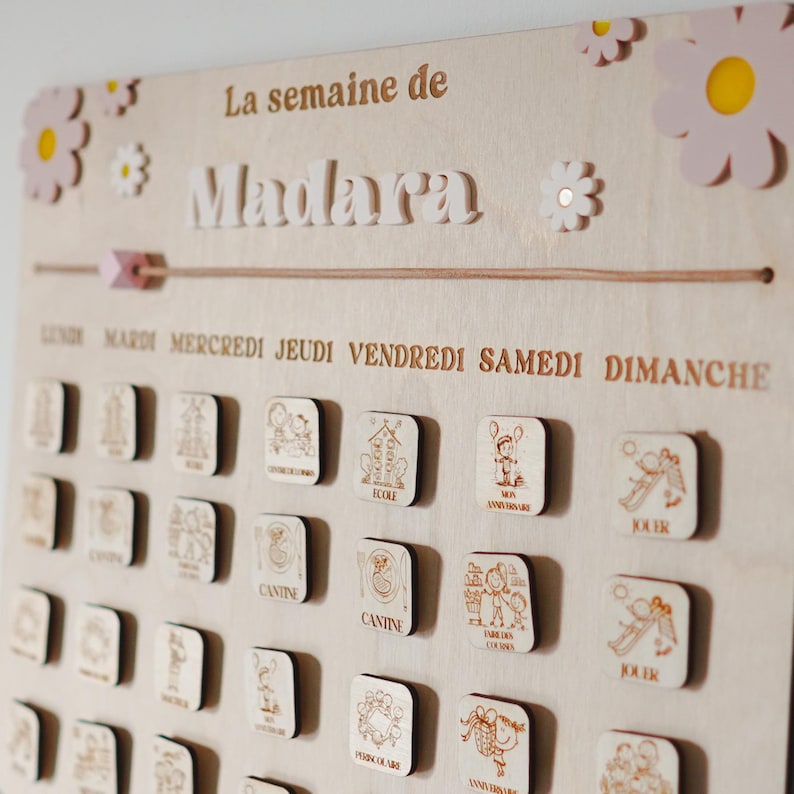 Pu&ograve; includere: Un calendario in legno con il nome Madara scritto su di esso. Il calendario ha i giorni della settimana scritti in francese e ha piccoli quadrati di legno con immagini di attivit&agrave; per ogni giorno. Il calendario &egrave; decorato con fiori rosa.