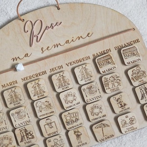 May include: A wooden calendar with a light brown finish. The calendar is designed for a child's weekly schedule. It has a title that says "ma semaine" which translates to "my week" in English. The calendar has days of the week labeled in French: Mardi, Mercredi, Jeudi, Vendredi, Samedi, Dimanche. Each day has a different image representing an activity, such as school, sports, or family time.