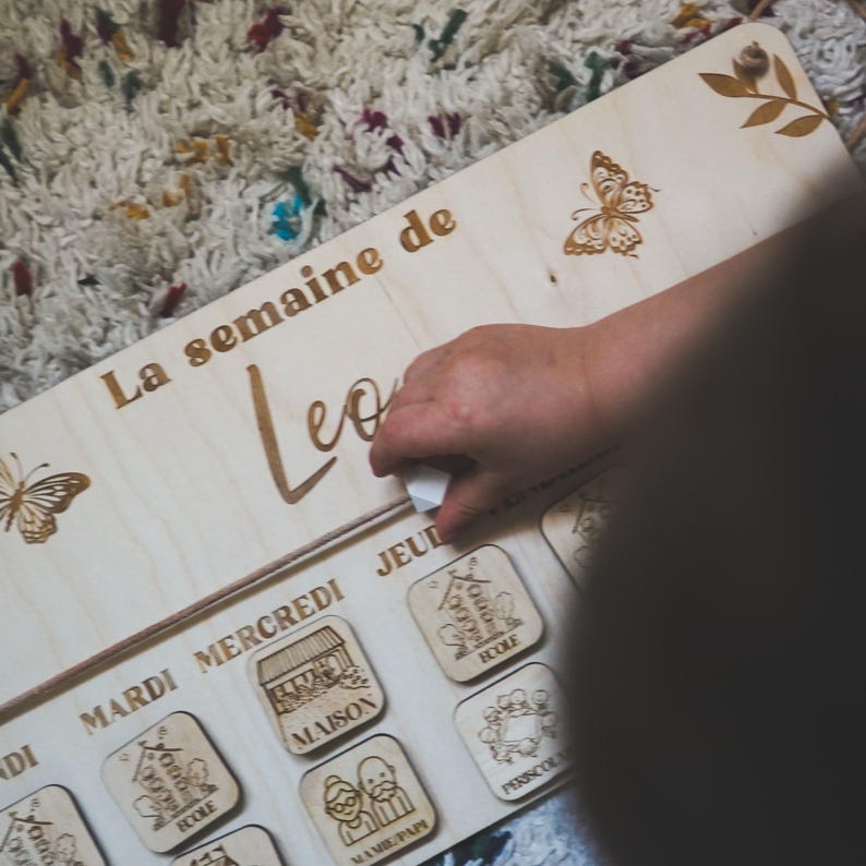 Pu&ograve; includere: Un pannello di legno con il testo "La semaine de Leo" e un design a farfalla. Il pannello ha una serie di piccoli quadrati di legno con immagini e testo, tra cui "Mardi", "Mercredi", "Jeu", "Ecole", "Maison", "Mamie Papi" e "Periscol".
