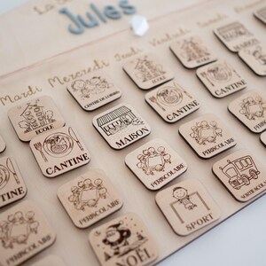 May include: Wooden children's schedule board with engraved icons and text. The board features the name "Jules" and days of the week. Square wooden tiles depict activities like "School", "Canteen", and "Sport".