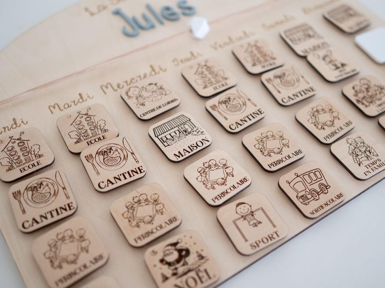 May include: Wooden children's schedule board with engraved icons and text. The board features the name "Jules" and days of the week. Square wooden tiles depict activities like "School", "Canteen", and "Sport".