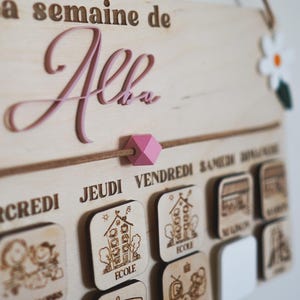 May include: A wooden weekly schedule board with the name "Alba" written in pink lettering. The board has days of the week labeled in French and includes small wooden icons for each day, such as a house for school, a bus for transportation, and a family for family time.