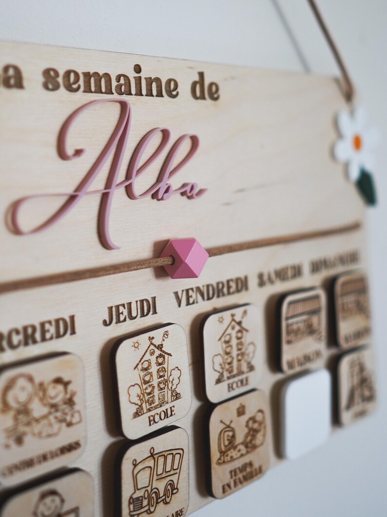 May include: A wooden weekly schedule board with the name "Alba" written in pink lettering. The board has days of the week labeled in French and includes small wooden icons for each day, such as a house for school, a bus for transportation, and a family for family time.