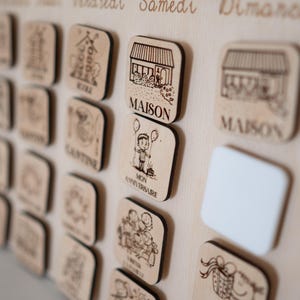 May include: Wooden calendar board with engraved icons and text. Includes square wooden tiles with illustrations of a house, a child with balloons, and other symbols. The board has the days of the week written in French.