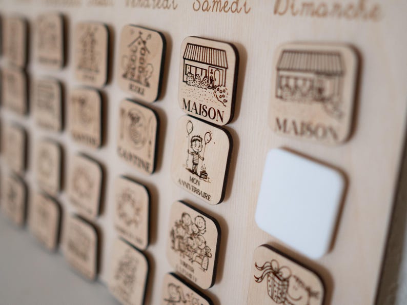 May include: Wooden calendar board with engraved icons and text. Includes square wooden tiles with illustrations of a house, a child with balloons, and other symbols. The board has the days of the week written in French.