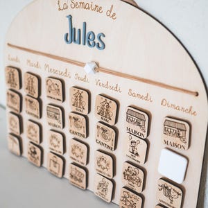 May include: Wooden weekly calendar with the text "La Semaine de Jules". The calendar has wooden square tiles with illustrations for each day of the week. The days of the week are written in French.
