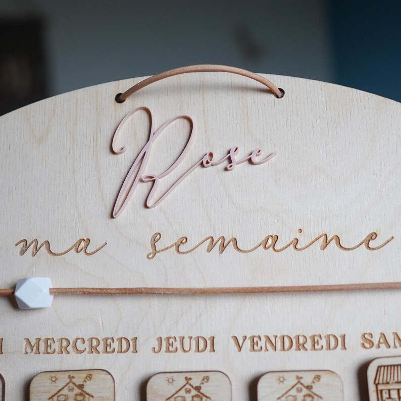 May include: A wooden board with the words "Rose ma semaine" engraved on it. The board has a pink, cursive "Rose" at the top and a wooden dowel with a white bead at the top. The board has days of the week written on it: "Mercredi, Jeudi, Vendredi, Samedi".