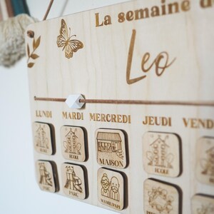Pu&ograve; includere: Un pannello di legno per la programmazione settimanale con il testo "La semaine de Leo" in alto. Il pannello presenta un design a farfalla ed &egrave; diviso in sette giorni della settimana: Lundi, Mardi, Mercredi, Jeudi, Vendredi, Samedi e Dimanche. Ogni giorno ha un piccolo quadrato di legno con un'immagine diversa, come un edificio scolastico, una casa, un parco e un'immagine dei nonni.