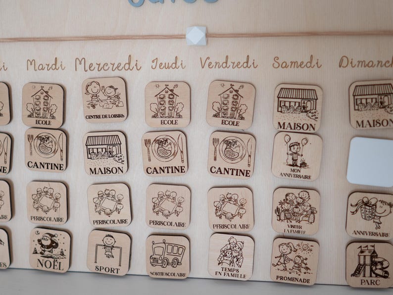 May include: Wooden calendar with engraved icons and French text. The calendar features various activity icons, including school, home, and dining, with the days of the week written across the top. The calendar is designed for educational or organizational purposes.