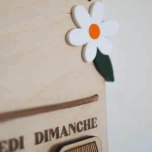 May include: A wooden calendar with the days of the week labeled in French. The days of the week are: Lundi, Mardi, Mercredi, Jeudi, Vendredi, Samedi, Dimanche. The calendar has a white daisy with an orange center and green leaves.