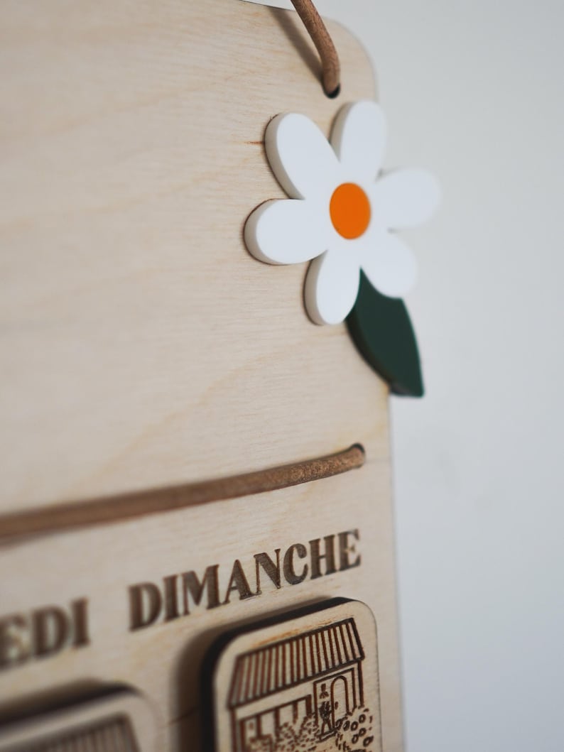 May include: A wooden calendar with the days of the week labeled in French. The days of the week are: Lundi, Mardi, Mercredi, Jeudi, Vendredi, Samedi, Dimanche. The calendar has a white daisy with an orange center and green leaves.