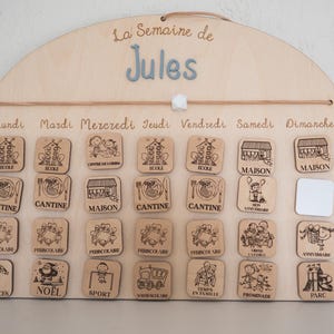 May include: Wooden weekly schedule board with the name "Jules" in blue letters. The board has a semi-circular top and includes wooden tiles with illustrations for each day of the week, such as school, home, and various activities.