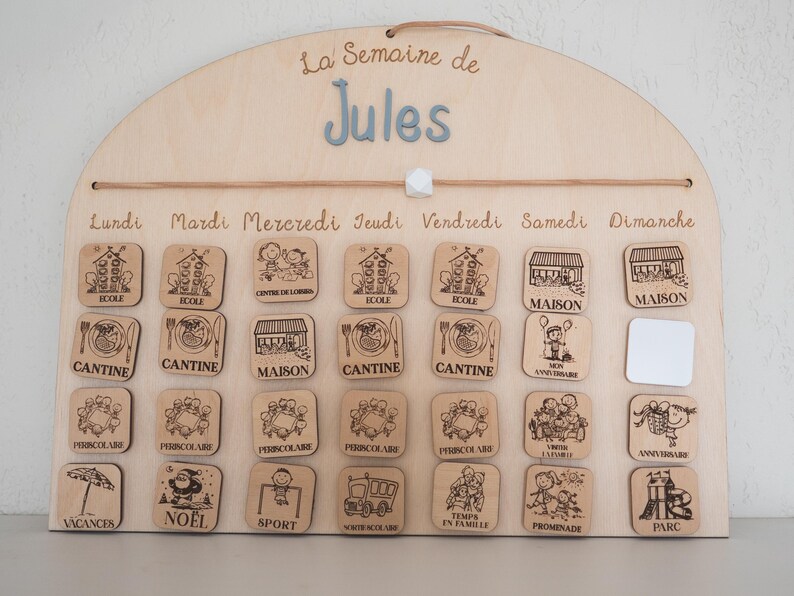 May include: Wooden weekly schedule board with the name "Jules" in blue letters. The board has a semi-circular top and includes wooden tiles with illustrations for each day of the week, such as school, home, and various activities.