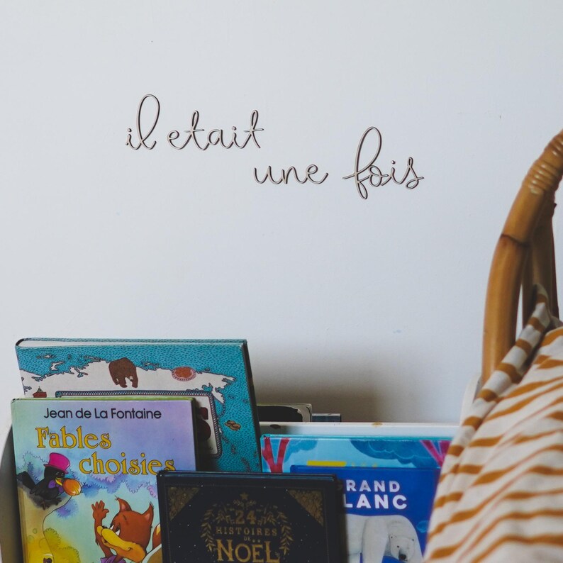 Op de afbeelding: Een witte muur met de Franse zin "il etait une fois" in cursief geschreven. Onder de tekst op de muur staat een stapel boeken op een plank. Het bovenste boek is blauw met een kaart op de omslag. Het tweede boek is roze met de titel "Fables Choisies" van Jean de la Fontaine. Het derde boek is zwart met gouden letters en de titel "24 Histoires de Noel". Het vierde boek is blauw met de titel "Grand Blanc".