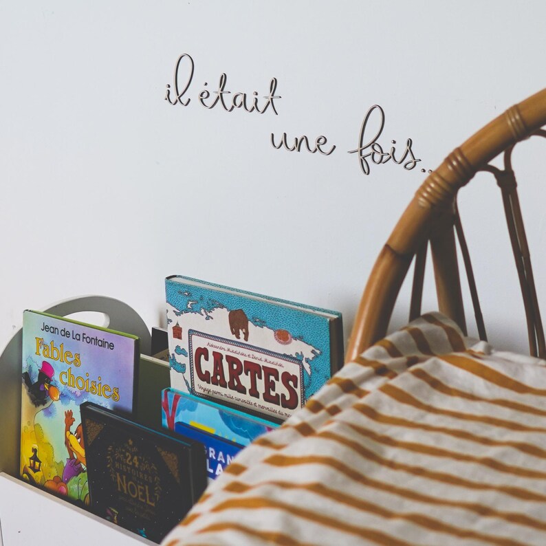 Op de afbeelding: Een witte boekenkast met een selectie kinderboeken, waaronder "Fables Choisies" van Jean de La Fontaine en een boek met de titel "Cartes". De boekenkast staat tegen een witte muur met de Franse zin "il &eacute;tait une fois" erop.