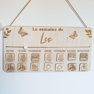 Pu&ograve; includere: Un pannello di programmazione settimanale in legno con il testo "La semaine de Leo" in alto. Il pannello ha un marcatore scorrevole e una fila di tessere di legno con immagini e testo per ogni giorno della settimana. Le tessere includono immagini di una casa, un parco e una scuola.