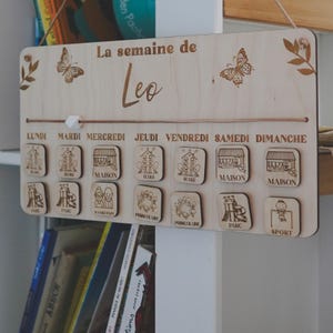 Pu&ograve; includere: Un pannello di programmazione settimanale in legno per un bambino di nome Leo. Il pannello ha i giorni della settimana etichettati in francese e include immagini di attivit&agrave; come la scuola, la casa, il parco e lo sport.
