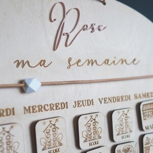 May include: A wooden weekly calendar with the words "Rose ma semaine" at the top. The calendar has days of the week labeled in French: "Lundi", "Mardi", "Mercredi", "Jeudi", "Vendredi", "Samedi", and "Dimanche". Each day has a small wooden square with a picture of a house and the word "ecole" on it.