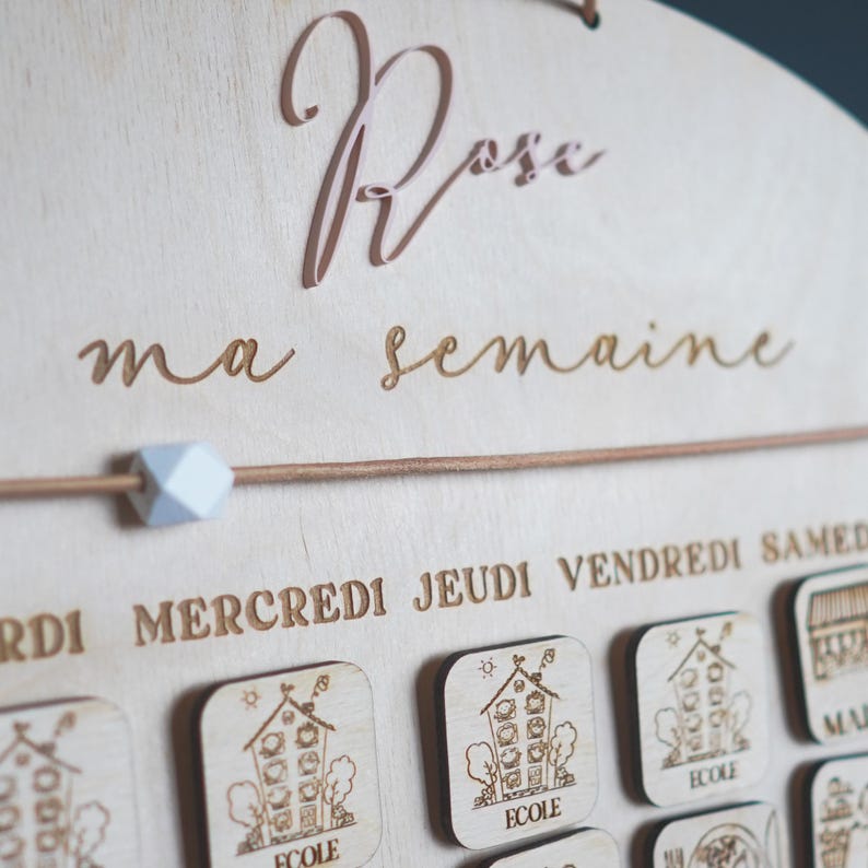 May include: A wooden weekly calendar with the words "Rose ma semaine" at the top. The calendar has days of the week labeled in French: "Lundi", "Mardi", "Mercredi", "Jeudi", "Vendredi", "Samedi", and "Dimanche". Each day has a small wooden square with a picture of a house and the word "ecole" on it.