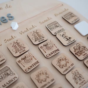 May include: Wooden weekly schedule board with engraved illustrations and text. Includes days of the week, the name "Jules", and various activity icons like school, home, and canteen. A decorative and functional item.