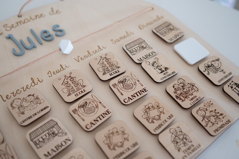 May include: Wooden weekly schedule board with engraved illustrations and text. Includes days of the week, the name "Jules", and various activity icons like school, home, and canteen. A decorative and functional item.