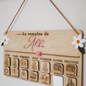 May include: A wooden weekly schedule board with the name "Alba" at the top. The board has seven days of the week labeled in French, with corresponding icons for each day. The icons include school, canteen, sports, school bus, family time, home, and park.