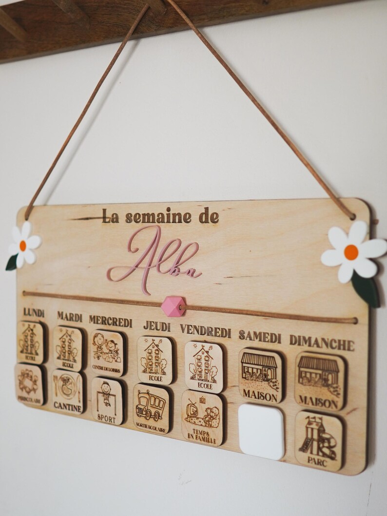 May include: A wooden weekly schedule board with the name "Alba" at the top. The board has seven days of the week labeled in French, with corresponding icons for each day. The icons include school, canteen, sports, school bus, family time, home, and park.