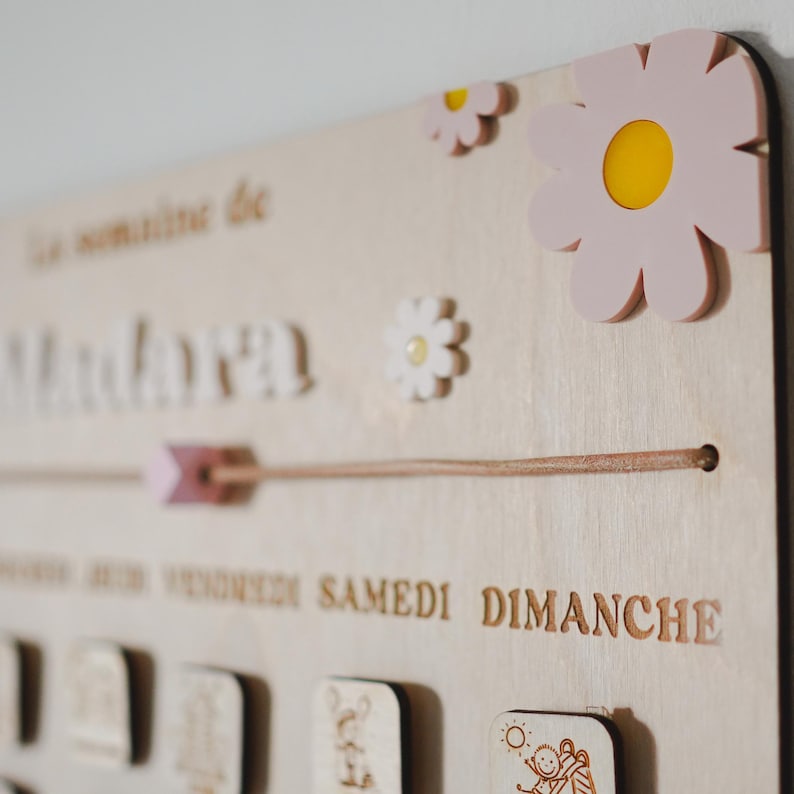 Pu&ograve; includere: Un calendario in legno con sfondo marrone chiaro e dettagli di fiori rosa e bianchi. Il calendario ha i giorni della settimana scritti in francese: Lundi, Mardi, Mercredi, Jeudi, Vendredi, Samedi, Dimanche.