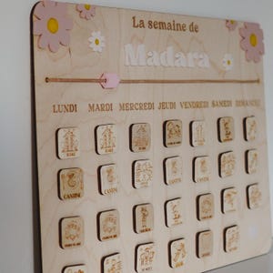 Pu&ograve; includere: Un pannello di programmazione settimanale in legno con il nome "Madara" in alto. Il pannello &egrave; decorato con fiori rosa e ha una corda con un'etichetta rosa. Il pannello ha i giorni della settimana etichettati in francese e include icone per attivit&agrave; come la scuola, la mensa e il tempo di gioco.