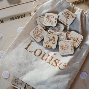 Pu&ograve; includere: Una borsa di tela bianca con lettere dorate scintillanti che dicono "Louise". La borsa contiene tessere quadrate di legno con illustrazioni e parole francesi, come "Jouer" e "Anniversaire".