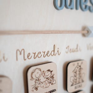 May include: Wooden wall calendar with engraved days of the week in French. Features small wooden squares with illustrations and the word "loisirs". The word "ouies" is in blue 3D letters.