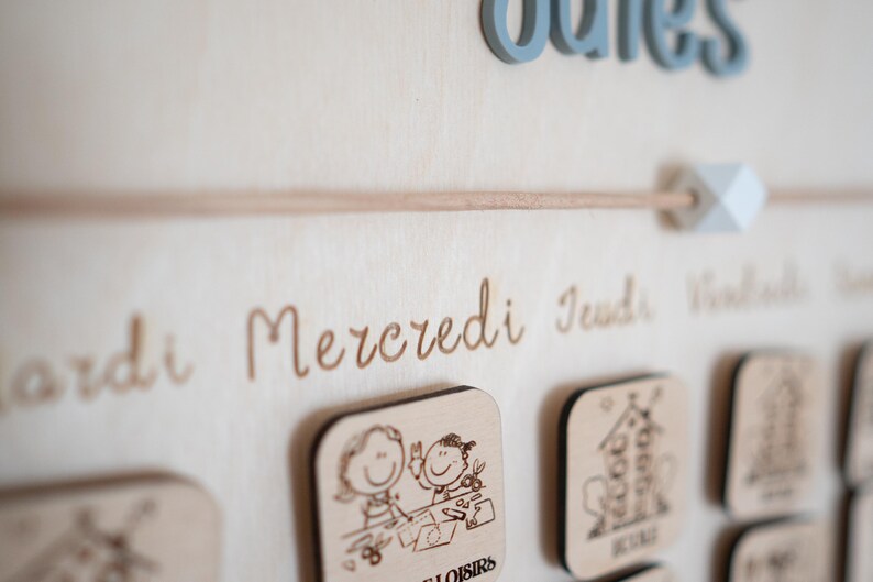 May include: Wooden wall calendar with engraved days of the week in French. Features small wooden squares with illustrations and the word "loisirs". The word "ouies" is in blue 3D letters.