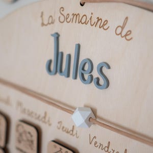 May include: Wooden weekly calendar with the name "Jules" in blue 3D letters. The text "La Semaine de" is above the name in a brown script font. A light brown string runs across the calendar with a white geometric bead.