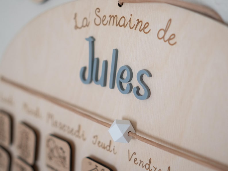 May include: Wooden weekly calendar with the name "Jules" in blue 3D letters. The text "La Semaine de" is above the name in a brown script font. A light brown string runs across the calendar with a white geometric bead.