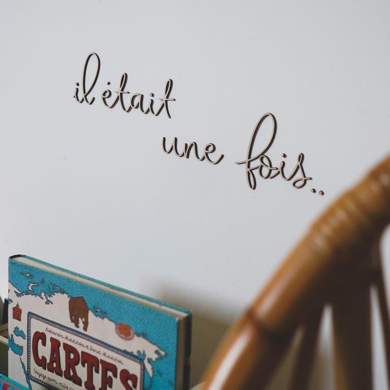 Op de afbeelding: Een witte muur met de Franse zin "il &eacute;tait une fois" geschreven in cursief schrift. De zin is gemaakt van hout en is bevestigd aan de muur. Een houten stoel bevindt zich op de voorgrond.