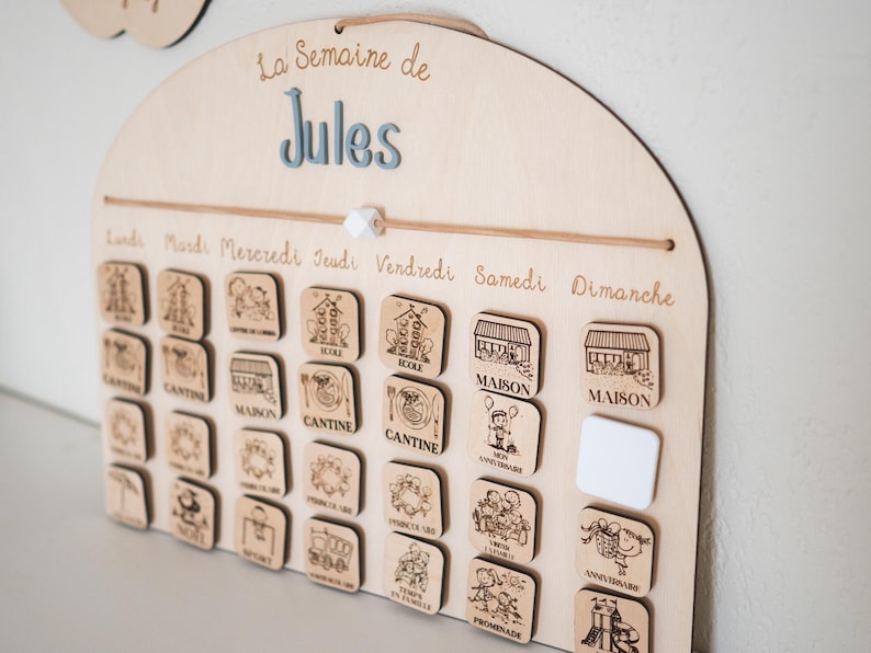 May include: Wooden weekly schedule board with the text "La Semaine de Jules". The board has a series of small wooden tiles with engraved illustrations and text, including "CANTINE" and "MAISON". The days of the week are labeled across the top.