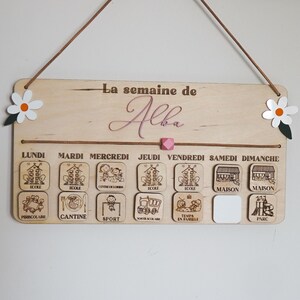 May include: A wooden weekly schedule board with the name "Alba" written in pink cursive. The board has seven days of the week labeled in French, with corresponding icons for each day. The icons include school, canteen, sports, after-school activities, family time, home, and park.
