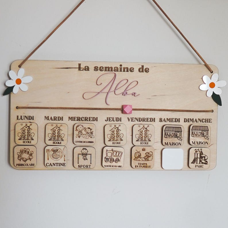 May include: A wooden weekly schedule board with the name "Alba" written in pink cursive. The board has seven days of the week labeled in French, with corresponding icons for each day. The icons include school, canteen, sports, after-school activities, family time, home, and park.
