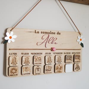 May include: A wooden weekly schedule board with the text "La semaine de Alba" at the top. The board has seven days of the week labeled in French, with corresponding icons for each day's activities, such as school, sports, and family time.
