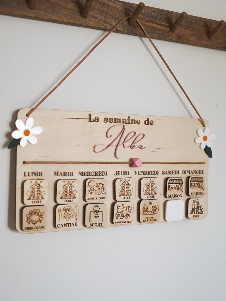 May include: A wooden weekly schedule board with the text "La semaine de Alba" at the top. The board has seven days of the week labeled in French, with corresponding icons for each day's activities, such as school, sports, and family time.