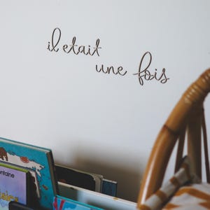 Op de afbeelding: Een witte muur met de Franse zin "il etait une fois" geschreven in cursief schrift. Op de voorgrond staat een houten stoel met een gestreept kussen.