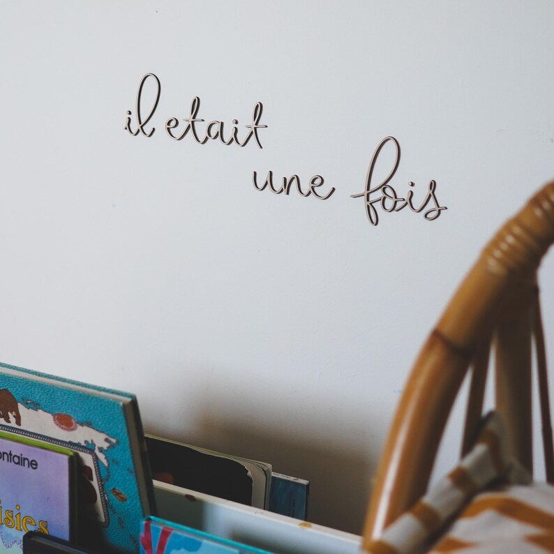 Op de afbeelding: Een witte muur met de Franse zin "il etait une fois" geschreven in cursief schrift. Op de voorgrond staat een houten stoel met een gestreept kussen.