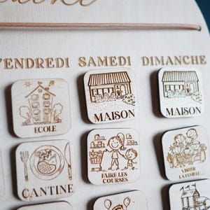 May include: Wooden calendar with magnets featuring illustrations of daily activities, including school, home, grocery shopping, and visiting family. The days of the week are labeled in French: Vendredi, Samedi, Dimanche.