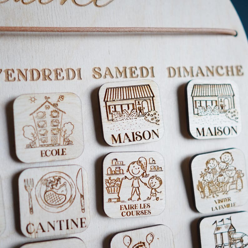 May include: Wooden calendar with magnets featuring illustrations of daily activities, including school, home, grocery shopping, and visiting family. The days of the week are labeled in French: Vendredi, Samedi, Dimanche.