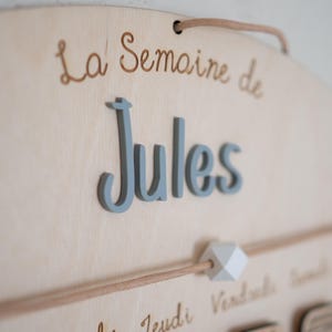 May include: Wooden wall hanging with the text "La Semaine de Jules" in script. The name "Jules" is in 3D gray letters. A wooden dowel with a white geometric bead is below the name.