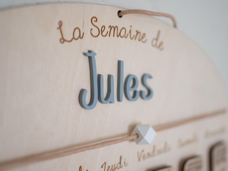 May include: Wooden wall hanging with the text "La Semaine de Jules" in script. The name "Jules" is in 3D gray letters. A wooden dowel with a white geometric bead is below the name.