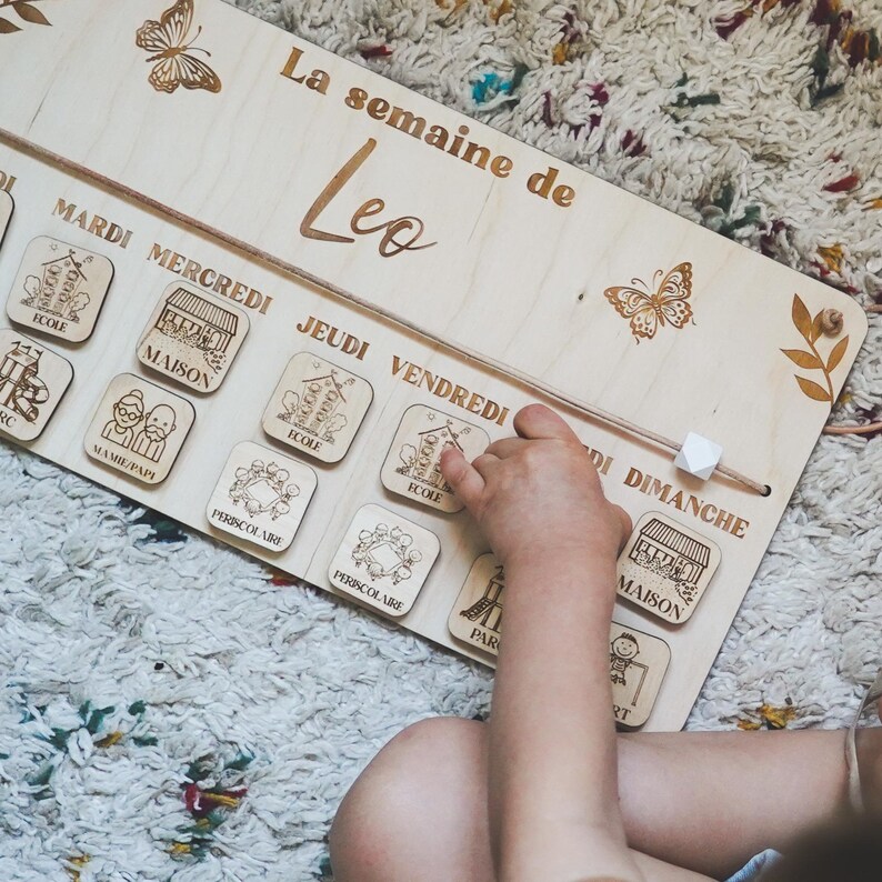 Pu&ograve; includere: Un pannello di programmazione settimanale in legno con il nome "Leo" in alto. Il pannello ha i giorni della settimana etichettati in francese e include immagini di attivit&agrave; come la scuola, la casa e il parco. La mano di un bambino sposta un cursore in legno per indicare il giorno corrente.